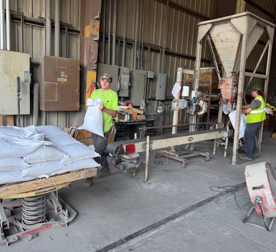Worker carrying a filled 50 lb bag beside stacked bags on a pallet lift, with a second worker at the hopper filling station.