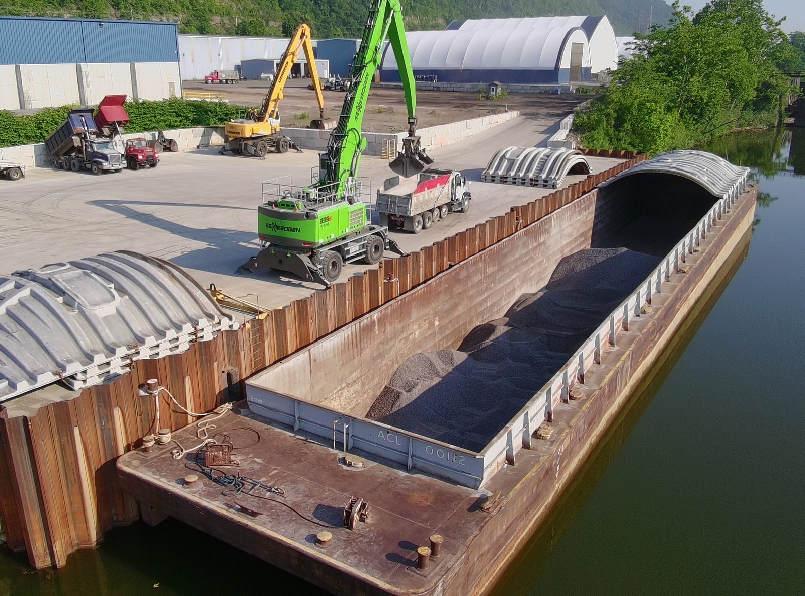 Green material handler loading a dump truck beside an open barge of bulk material at the terminal dock.