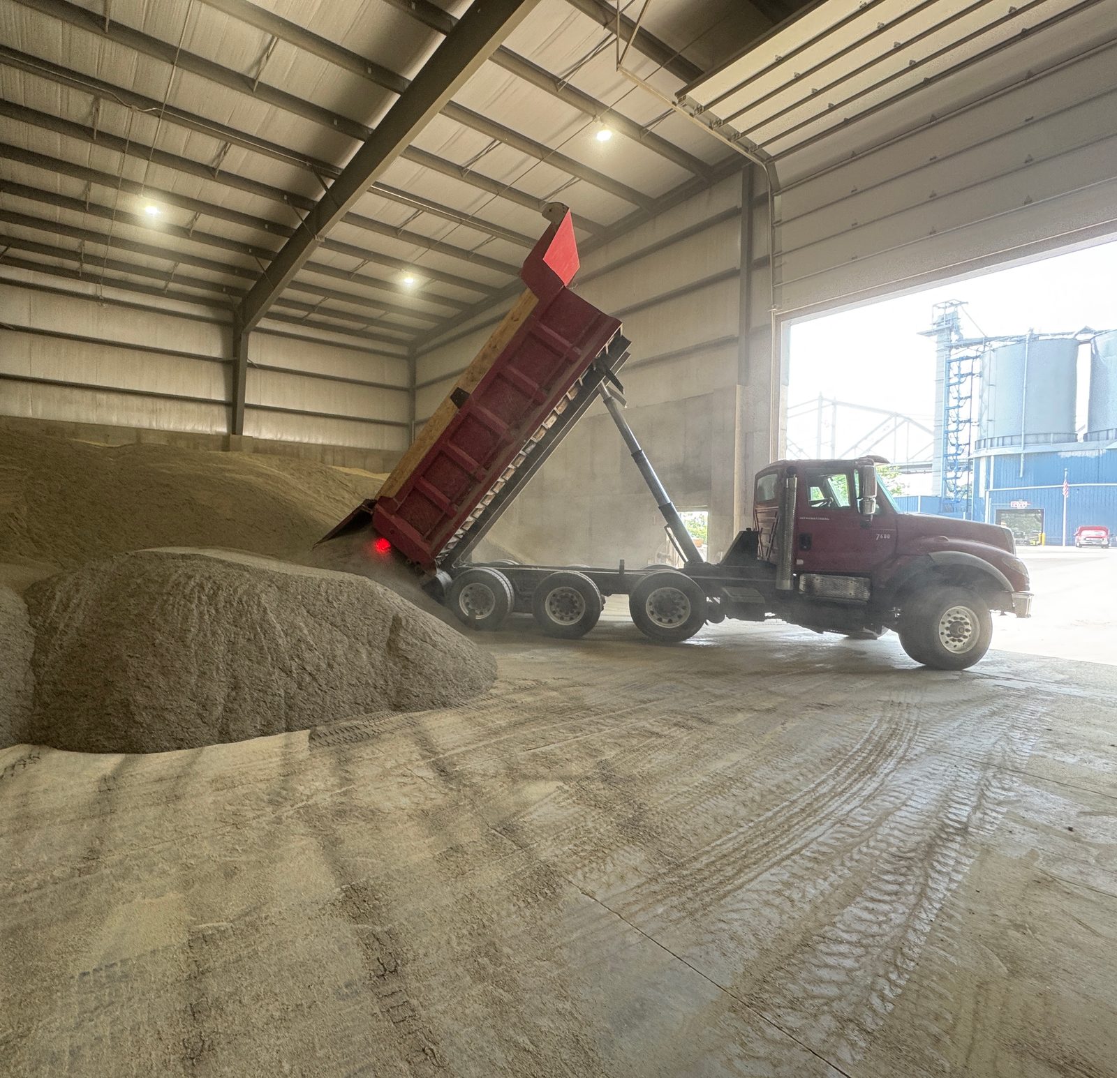 Dump truck with its bed raised unloading bulk material onto a pile inside a warehouse, silos visible through the doorway.
