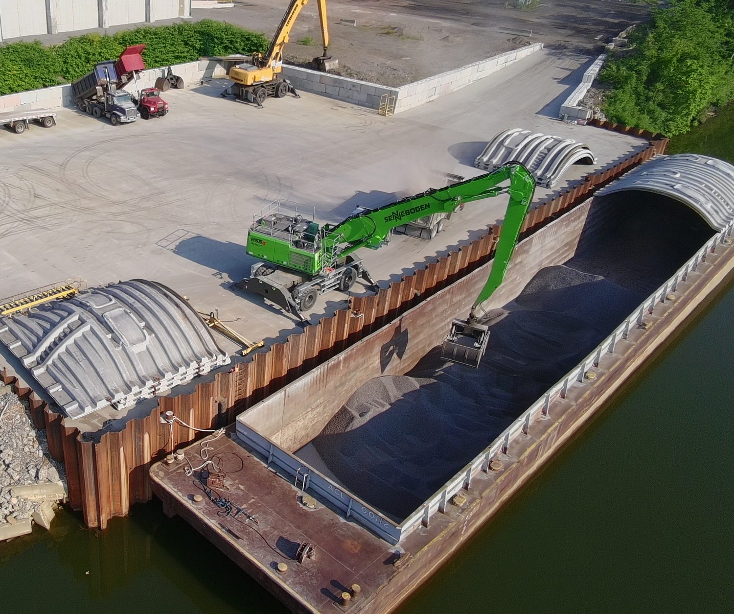 Material handler on the dock reaching its grab into an open barge of bulk material, with barge covers stacked alongside.