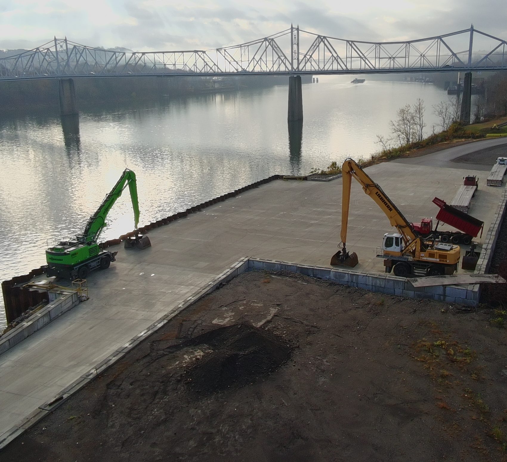 Green and yellow material handlers working on the concrete barge dock, with the Ohio River and a truss bridge beyond.