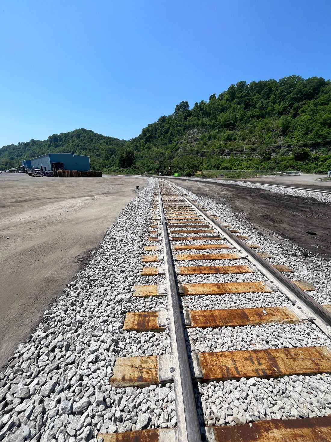 Rail line running through the terminal yard toward a wooded hillside, with a warehouse and trailers on the left.