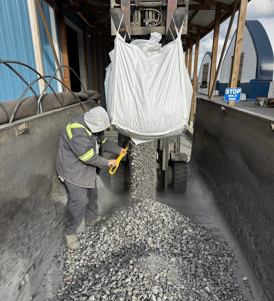 Worker emptying a super sack suspended from a forklift into a truck bed, with material pouring onto the pile below.