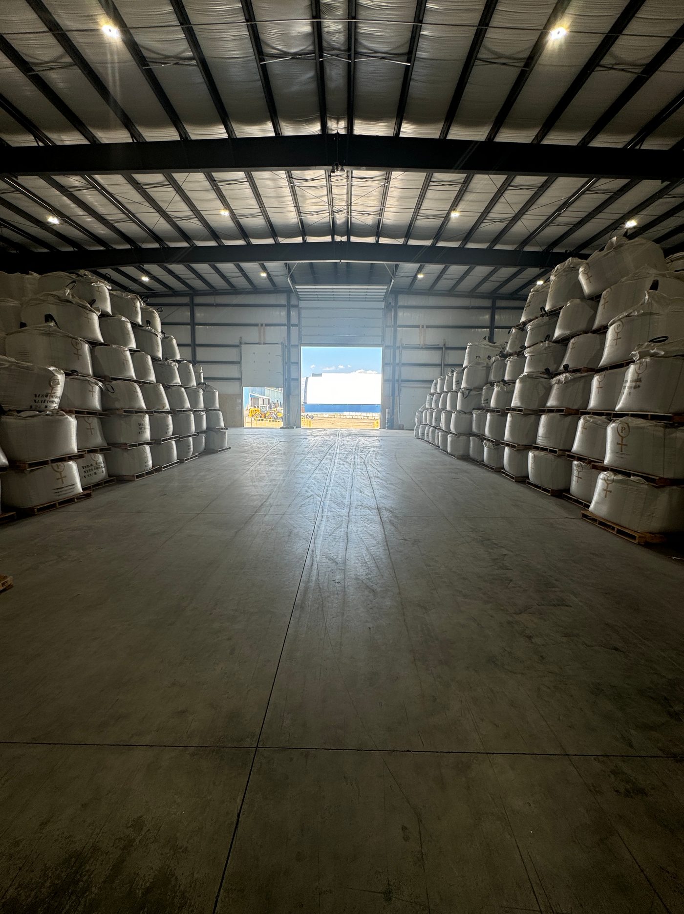 Rows of stacked super sacks lining both sides of a warehouse aisle, with a bright doorway at the far end.